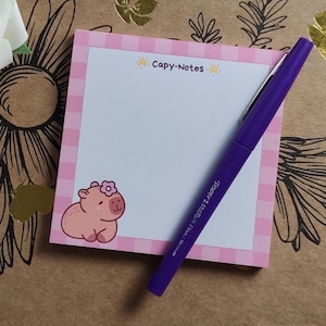 May include: A square notepad with a pink gingham border and a cartoon capybara illustration. The notepad has the text "Capy-Notes" and a purple Paper Mate pen is resting on it.