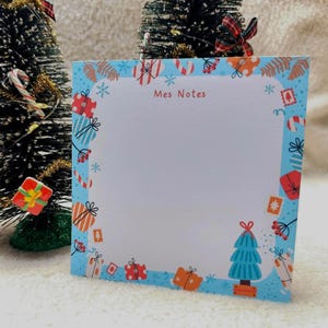 Peut inclure: Un bloc-notes bleu avec une bordure sur le thème de Noël, ornée de boîtes cadeaux, de flocons de neige et d'un sapin de Noël. Le bloc-notes porte l'inscription "Mes Notes". Le bloc-notes est debout.
