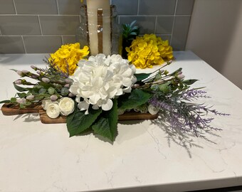 May include: A white hydrangea flower arrangement with white roses and purple lavender sprigs on a wooden tray. The arrangement is on a white countertop.
