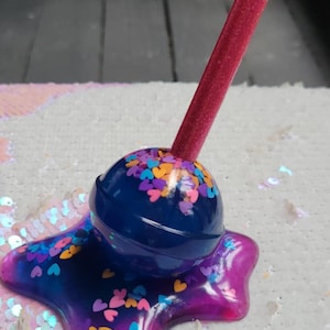 May include: A whimsical, handmade lollipop-shaped resin art piece. The dark blue candy has colorful heart-shaped sprinkles. The base is a vibrant mix of purple and blue resin. A magenta stick completes the design.