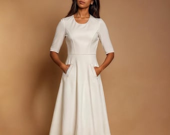 Puede incluir: Un vestido blanco hasta la rodilla con cuello redondo y mangas 3/4. El vestido tiene un corpiño ajustado y una falda acampanada con bolsillos.