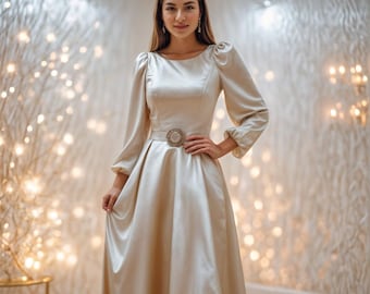 Op de afbeelding: Een vrouw draagt een lange, vloeiende, champagnekleurige satijnen jurk met een getailleerd lijfje en een volle rok. De jurk heeft een decoratieve riem met een gesp met juwelen. De vrouw staat voor een muur met een glinsterende achtergrond.