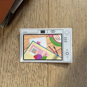Op de afbeelding: Een rechthoekige sticker in de vorm van een vintage televisie met een kleurrijke illustratie van een bureau. Op het scherm is een notitieboek, een witte envelop, een kop koffie, twee pennen, een aardbei en een kleine roze strik te zien.