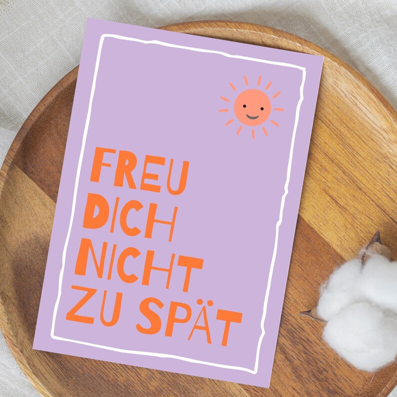 May include: A rectangular greeting card with a white border and a light purple background. The card features the German text "FREU DICH NICHT ZU SP&Auml;T" in orange, along with a smiling sun illustration. The card is placed on a wooden tray.