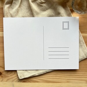May include: A blank white postcard with a stamp design in the upper right corner and horizontal lines for the address. The postcard is resting on a beige cloth, with a wooden surface in the background.