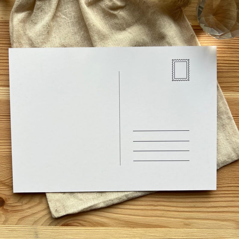 May include: A blank white postcard with a stamp design in the upper right corner and horizontal lines for the address. The postcard is resting on a beige cloth, with a wooden surface in the background.
