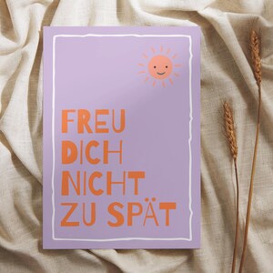 May include: A lilac-colored art print with a white border features the German text "FREU DICH NICHT ZU SP&Auml;T" in orange. A smiling sun graphic is in the upper right corner. The print is displayed on a beige fabric background.