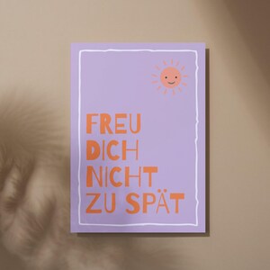 May include: A rectangular poster with a light purple background and a white border. The poster features the text "FREU DICH NICHT ZU SP&Auml;T" in orange, along with a smiling sun graphic in the upper right corner. The poster is displayed on a neutral-toned wall.