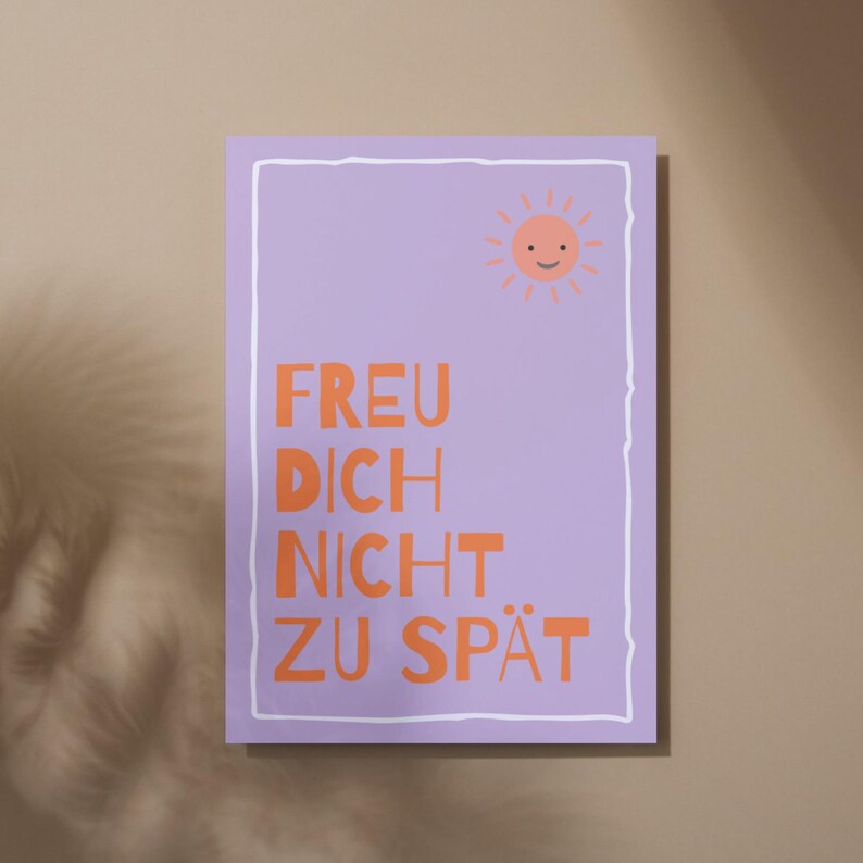 May include: A rectangular poster with a light purple background and a white border. The poster features the text "FREU DICH NICHT ZU SP&Auml;T" in orange, along with a smiling sun graphic in the upper right corner. The poster is displayed on a neutral-toned wall.
