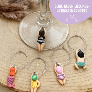 Könnte beinhalten: Weinmarkierer mit Miniaturfiguren in Badebekleidung. Die Charms umfassen eine Meerjungfrau, eine Figur in einem gelben Badeanzug und Figuren in blauen, rosa und schwarzen Badeanzügen. Die Charms sind an silbernen Reifen befestigt.