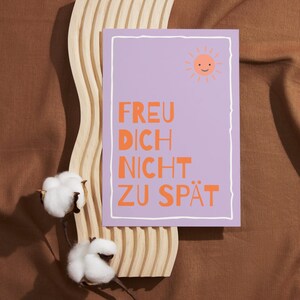 May include: A lilac-colored card with the text "FREU DICH NICHT ZU SP&Auml;T" in orange, and a smiling sun illustration. The card is set against a wooden decorative piece and brown fabric, with cotton sprigs.
