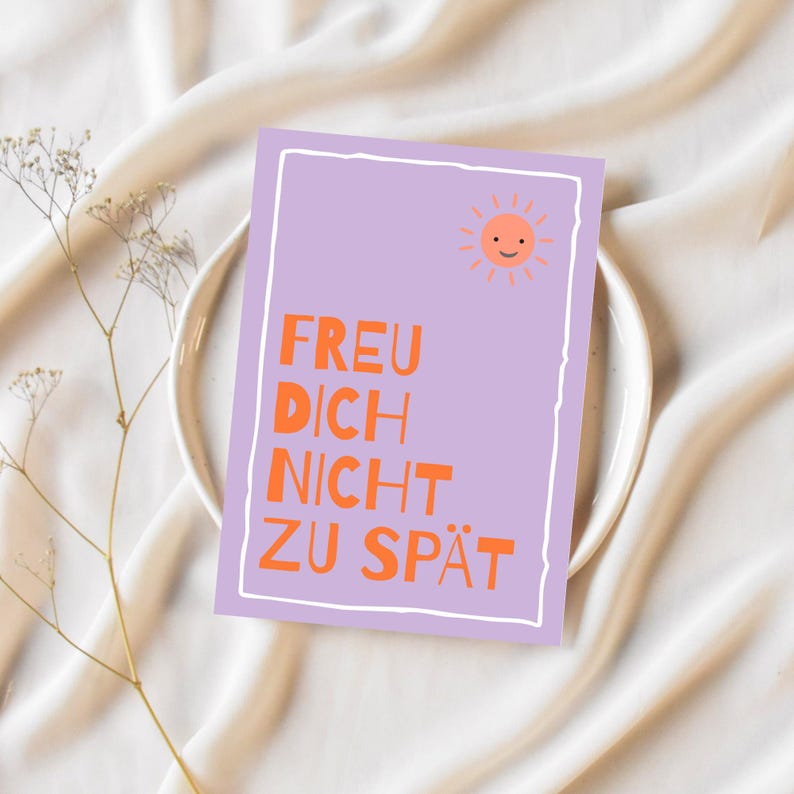 May include: A rectangular card with the text "FREU DICH NICHT ZU SP&Auml;T" in orange letters on a lavender background. A smiling sun graphic is in the upper right corner. The card is on a white plate, with a sprig of dried flowers to the left.