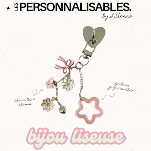 Puede incluir: Un llavero decorativo con un cierre en forma de corazón y una cadena con dijes. Los dijes incluyen un lazo rosa, una mariposa, flores de margarita, un corazón y una estrella rosa. El texto "LES PERSONNALISABLES" y "bijou liseuse" es visible.