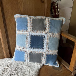 Könnte beinhalten: Quadratisches Zierkissen mit Patchwork-Design. Das Kissen hat Jeansstoffquadrate in verschiedenen Blau- und Grautönen, die mit einem hellblauen Häkelrand zusammengenäht sind. Das Kissen liegt auf einer Holzbank mit einem Kunstfell.