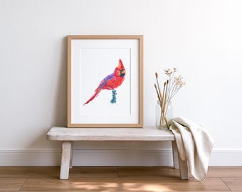 May include: A framed watercolor painting of a red cardinal bird with blue and purple accents. The artwork is displayed on a rustic wooden bench, alongside paintbrushes in a glass jar and a folded white cloth. The background is a white wall.