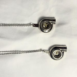 May include: Two silver necklaces with a turbocharger pendant. The pendants are detailed with a silver finish and a gold centre.