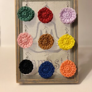 May include: A display of eight crocheted earrings in various colors, including green, red, purple, pink, brown, yellow, black, and orange. The earrings are displayed on a wooden frame with a wire mesh background.