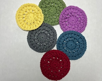Op de afbeelding: Zes gehaakte onderzetters in verschillende kleuren, waaronder geel, groen, paars, grijs, blauw en rood. De onderzetters zijn rond en hebben een textuur.