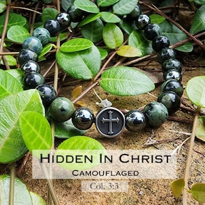 May include: A beaded bracelet featuring dark green and black stones, interspersed with metallic beads. A silver-toned circular charm with a cross design is centered on the bracelet. The text "HIDDEN IN CHRIST" and "CAMOUFLAGED" is visible.