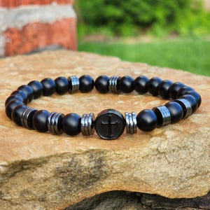 Puede incluir: Una pulsera elástica con cuentas esféricas de color negro mate intercaladas con espaciadores cilíndricos de color gris oscuro metálico. Una cuenta central circular negra muestra un símbolo de cruz en relieve, creando un diseño uniforme.
