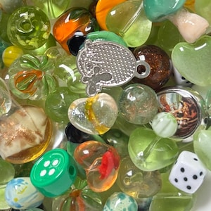 Puede incluir: Una colección de cuentas variadas en diversas formas, tamaños y colores. Las cuentas incluyen cuentas de vidrio verdes, transparentes y blancas, junto con algunas con patrones coloridos. Algunas cuentas tienen forma de corazón, otras son redondas o irregulares.