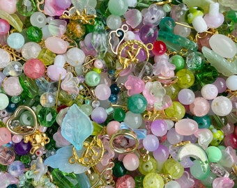 May include: A large assortment of colorful beads and charms in various shapes and sizes. The beads are mostly green, pink, and purple, with some clear and white beads. There are also gold charms in the shape of leaves, butterflies, and other designs.