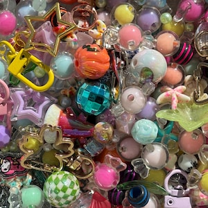 Può includere: Una collezione di perline artigianali e portachiavi in metallo assortiti in varie forme, dimensioni e colori. Il mix include sfere traslucide, ciondoli a forma di stella, perline a zucca arancioni, una palla da discoteca e perline rosa.