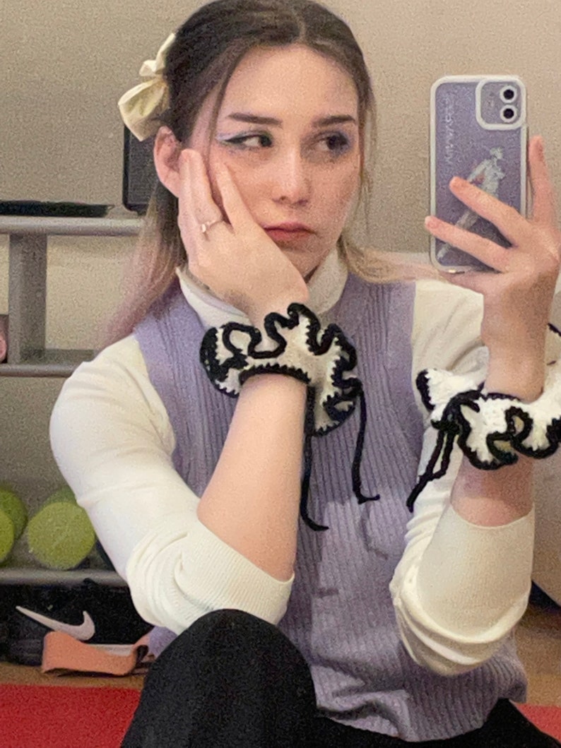 May include: A woman wearing a light purple sleeveless sweater with a white long-sleeved shirt underneath. She is wearing black and white crocheted bracelets on both wrists. She has a white bow in her hair and is holding a white phone case with a design on it.