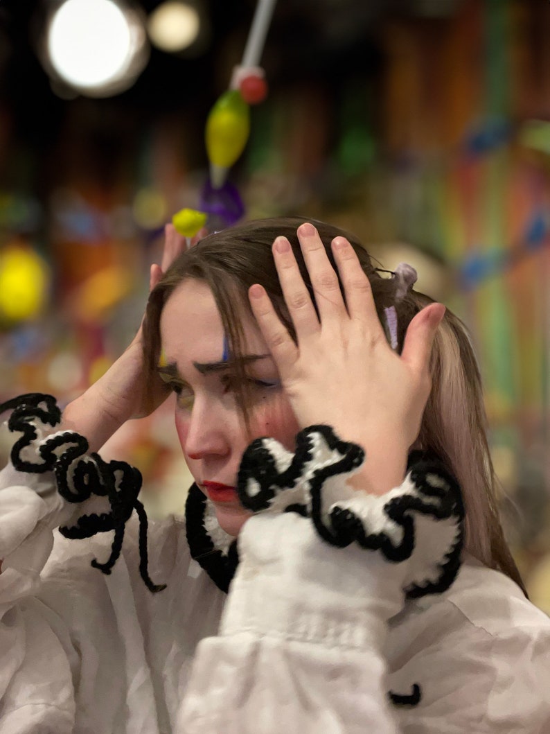 May include: A young person wearing a white shirt with black and white crocheted trim around the collar and cuffs. They have clown makeup on their face and are covering their face with their hands.