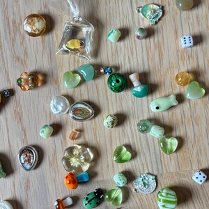 May include: A collection of small, colorful glass beads and charms scattered on a wooden surface. The assortment includes heart-shaped, flower, and fish-shaped beads, along with a miniature fish in a bag, dice, and other decorative elements.