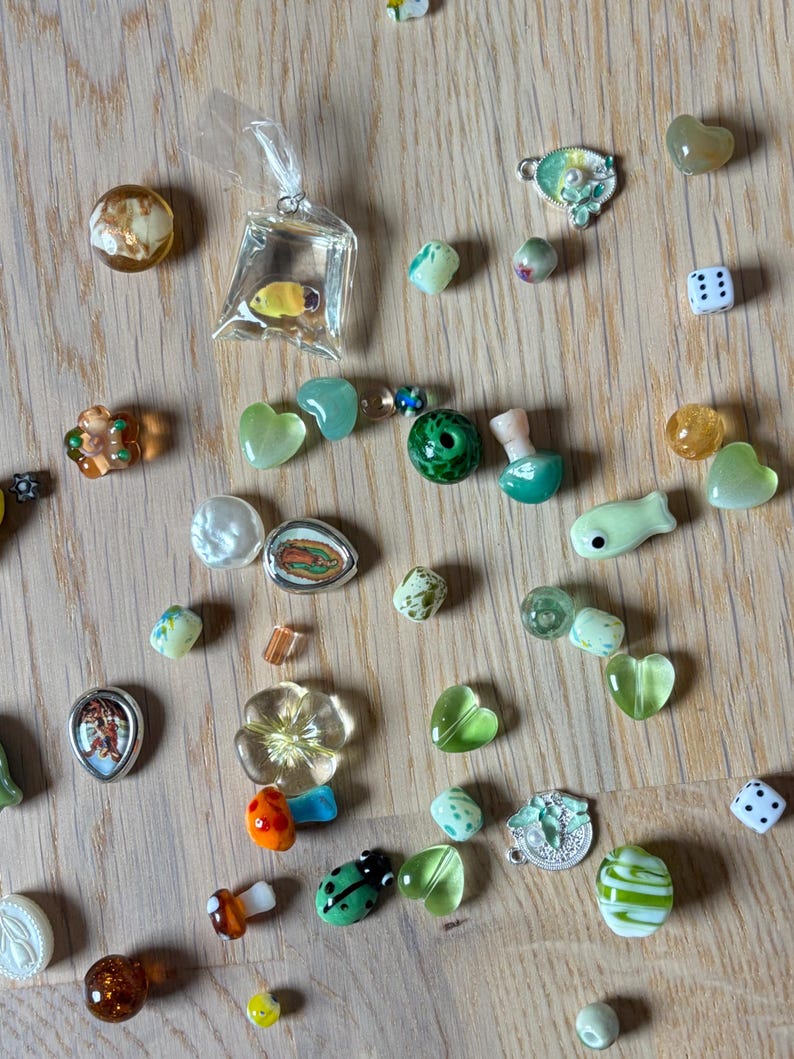 May include: A collection of small, colorful glass beads and charms scattered on a wooden surface. The assortment includes heart-shaped, flower, and fish-shaped beads, along with a miniature fish in a bag, dice, and other decorative elements.