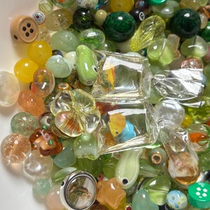 May include: A collection of colorful glass beads in various shapes and sizes. The beads include round, oval, and button-shaped designs, with colors ranging from green and yellow to clear and orange. Some beads have internal designs, and others are encased in clear glass.