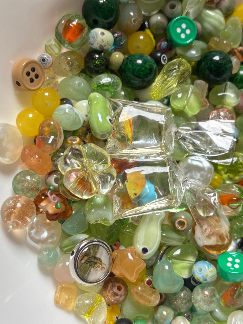 May include: A collection of colorful glass beads in various shapes and sizes. The beads include round, oval, and button-shaped designs, with colors ranging from green and yellow to clear and orange. Some beads have internal designs, and others are encased in clear glass.
