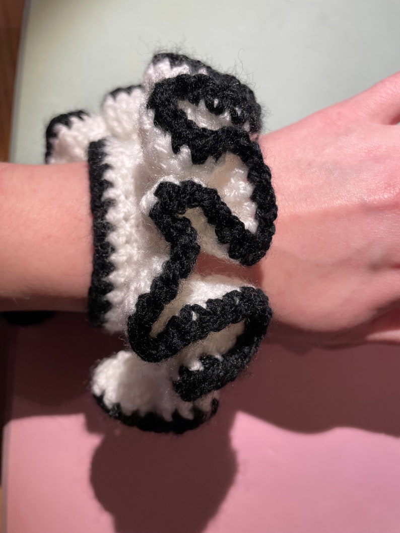 May include: White crocheted hair scrunchie with black trim. The scrunchie is gathered in a ruffled style.