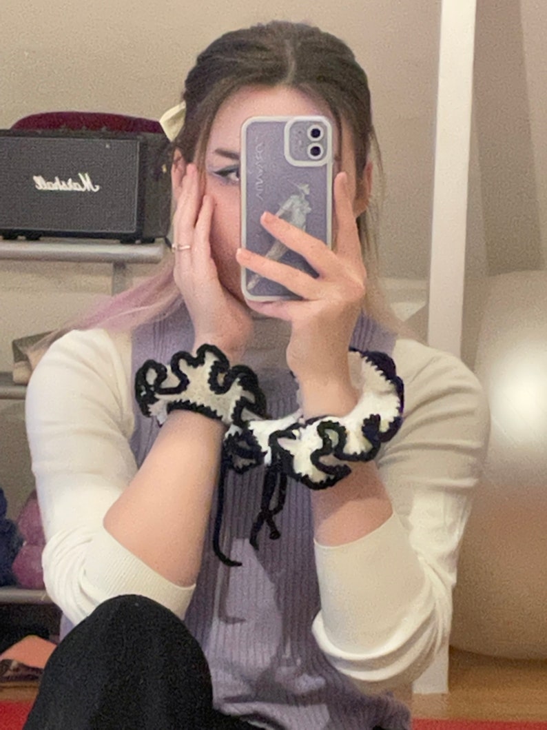May include: A person wearing a white and purple sweater with black and white crocheted bracelets. The person is holding a purple phone case with a white outline and a graphic of a person on it.