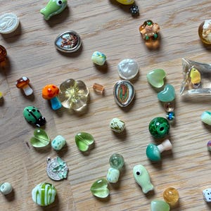 May include: A collection of colorful glass beads in various shapes, including fish, hearts, flowers, and mushrooms. The beads are in shades of green, yellow, orange, and white, with some featuring intricate designs and details. A few dice are also present.