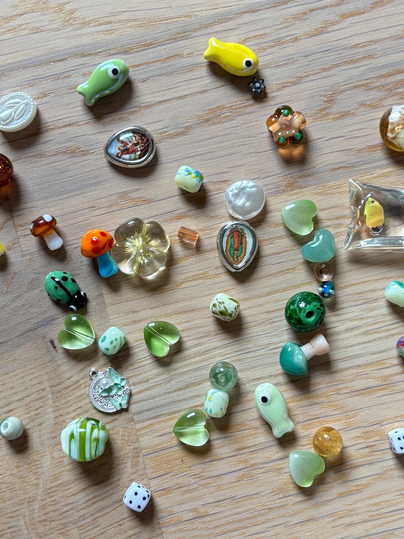 May include: A collection of colorful glass beads in various shapes, including fish, hearts, flowers, and mushrooms. The beads are in shades of green, yellow, orange, and white, with some featuring intricate designs and details. A few dice are also present.