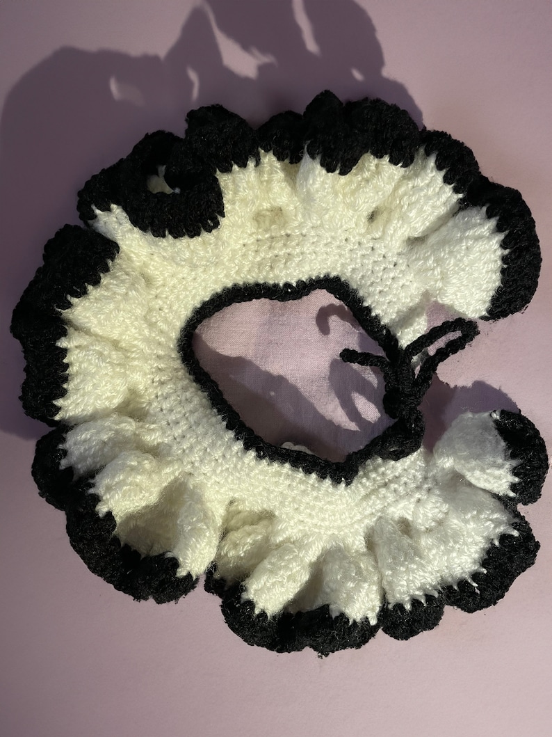 May include: A white crocheted collar with a black crocheted trim. The collar has a ruffled edge and a tie closure.