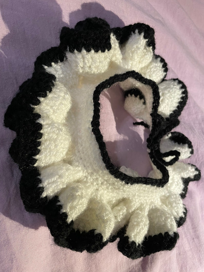 May include: A white crocheted collar with a black crocheted trim. The collar has a ruffled edge and is tied at the back with a black ribbon.