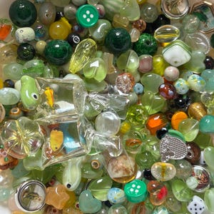 May include: A collection of assorted beads in shades of green, yellow, and clear. The beads vary in shape and size, including round, oval, and faceted designs. Some beads feature embedded designs, while others are solid colors. There are also buttons and small charms.