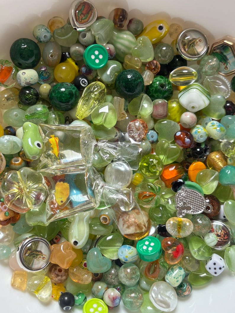 May include: A collection of assorted beads in shades of green, yellow, and clear. The beads vary in shape and size, including round, oval, and faceted designs. Some beads feature embedded designs, while others are solid colors. There are also buttons and small charms.