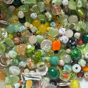 May include: A collection of assorted beads in various shapes, sizes, and colors. The beads include clear, opaque, and iridescent varieties, with shades of green, yellow, and brown. Some beads are heart-shaped, while others are round, oval, or square. There are also dice and other decorative elements.