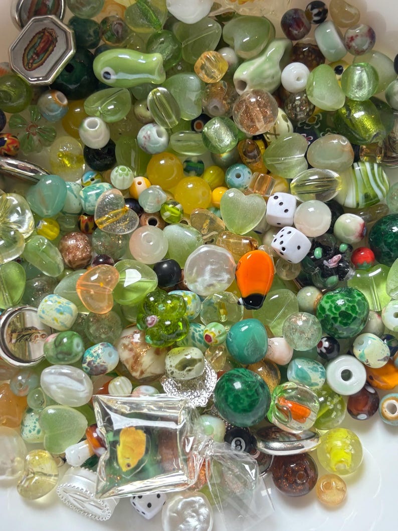May include: A collection of assorted beads in various shapes, sizes, and colors. The beads include clear, opaque, and iridescent varieties, with shades of green, yellow, and brown. Some beads are heart-shaped, while others are round, oval, or square. There are also dice and other decorative elements.