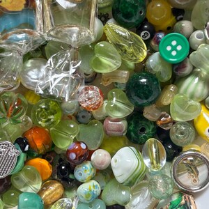 May include: A collection of various glass beads in shades of green, clear, and other colors. The beads include round, heart-shaped, and other unique designs. Some beads have embedded designs, and there is a green button with white dots.