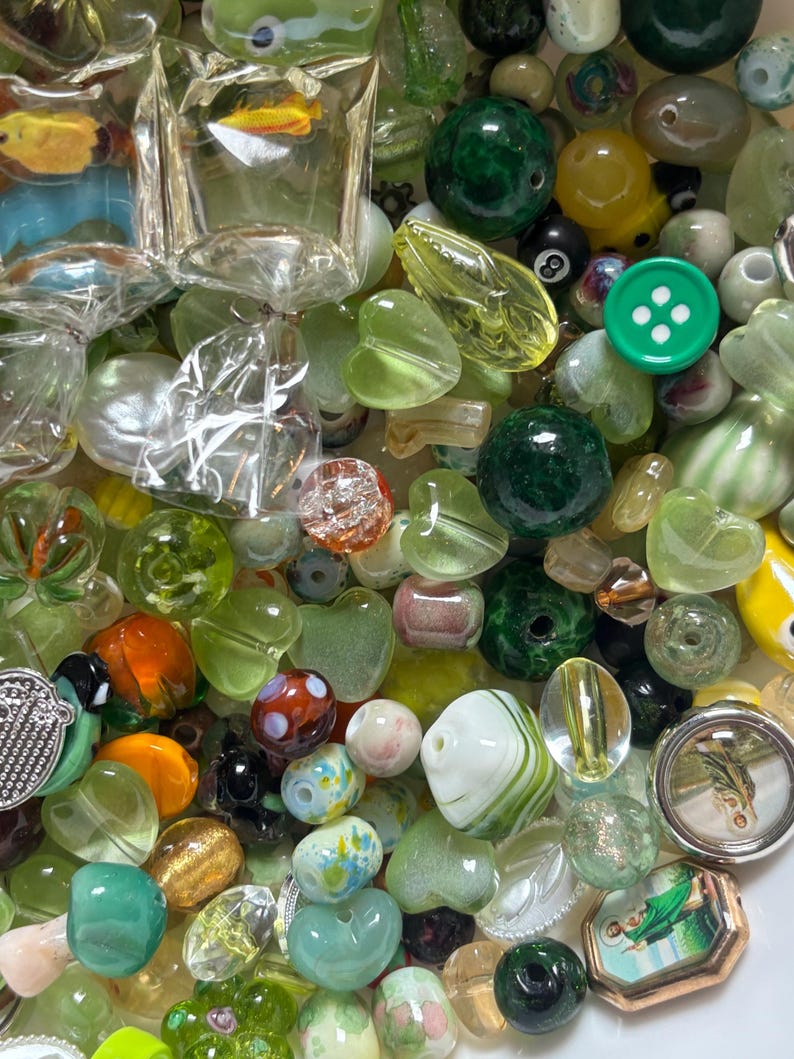 May include: A collection of various glass beads in shades of green, clear, and other colors. The beads include round, heart-shaped, and other unique designs. Some beads have embedded designs, and there is a green button with white dots.