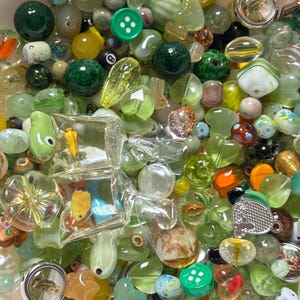 May include: A collection of various beads in shades of green, yellow, and clear. The assortment includes round, oval, and uniquely shaped beads, some with internal designs. A green button and a small die are also visible.