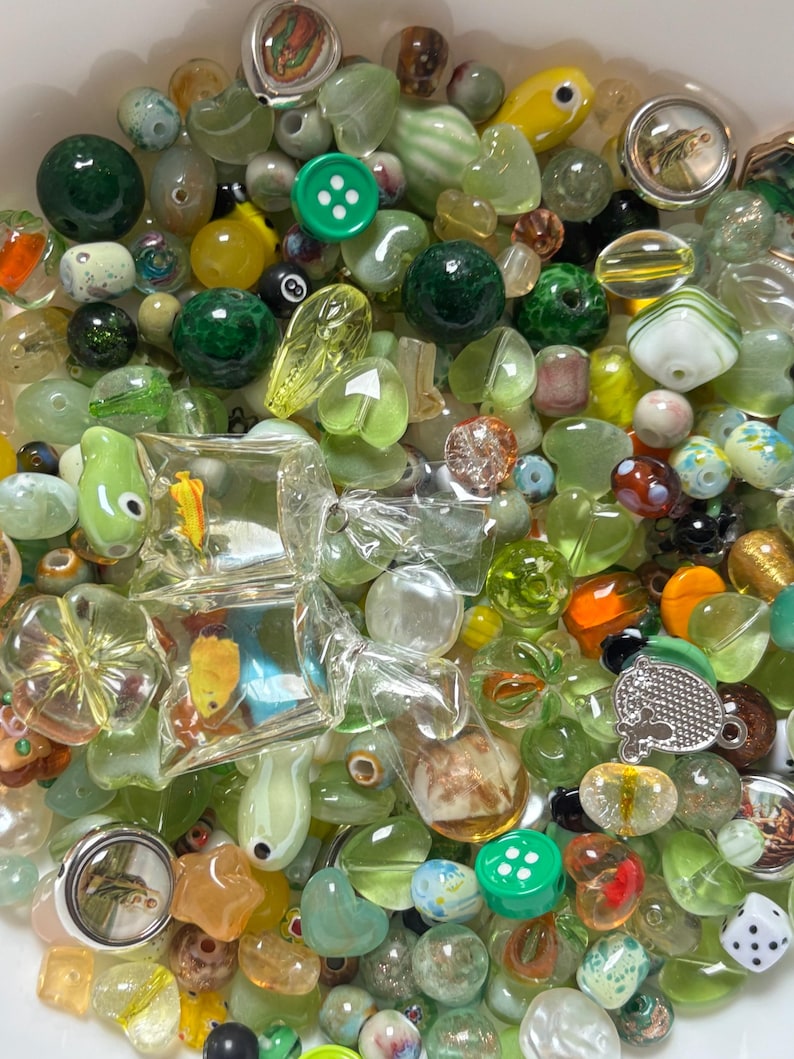May include: A collection of various beads in shades of green, yellow, and clear. The assortment includes round, oval, and uniquely shaped beads, some with internal designs. A green button and a small die are also visible.
