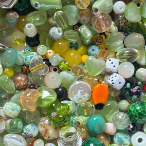 May include: A collection of colorful glass beads in various shapes and sizes. The beads are primarily green, yellow, and clear, with some featuring heart, dice, and floral designs. Some beads have a metallic sheen, and others are opaque.