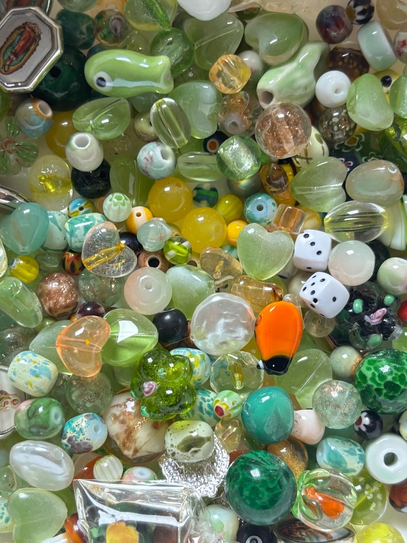 May include: A collection of colorful glass beads in various shapes and sizes. The beads are primarily green, yellow, and clear, with some featuring heart, dice, and floral designs. Some beads have a metallic sheen, and others are opaque.