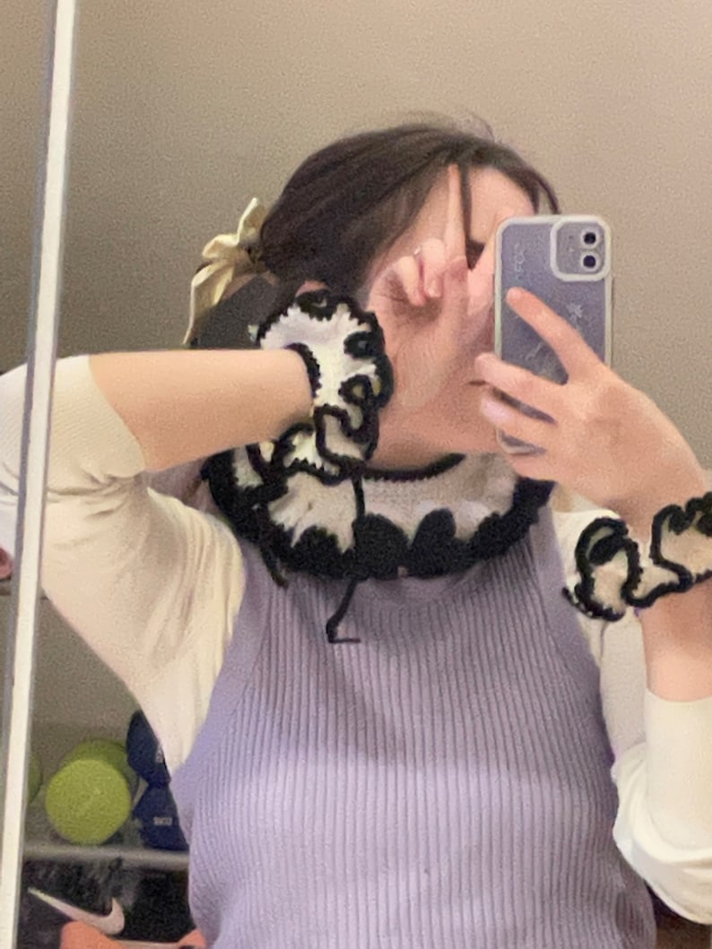 May include: A person wearing a purple sleeveless sweater with a black and white crocheted collar and matching bracelets. The person is wearing a white long-sleeved shirt underneath the sweater.